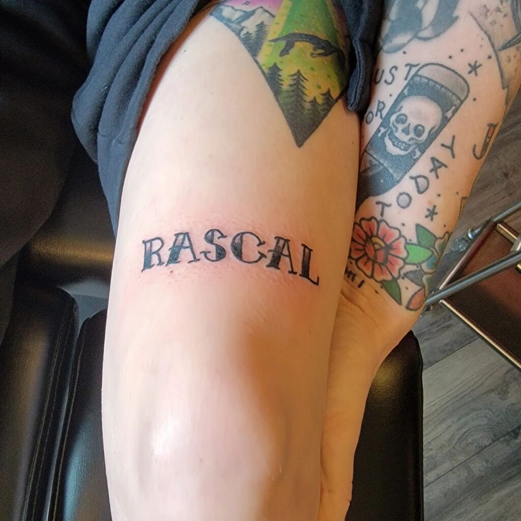 A playful word tattoo rascal above the knee