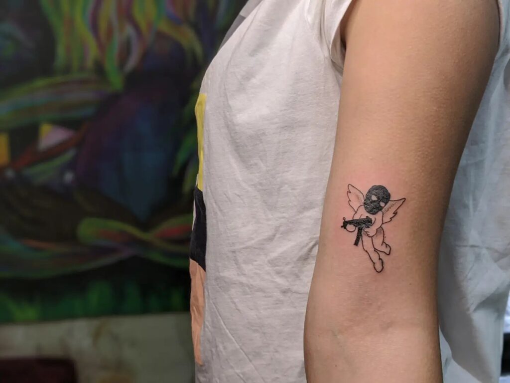 Above the elbow minimalist angel with gun tattoo symbolism with a face mask accent