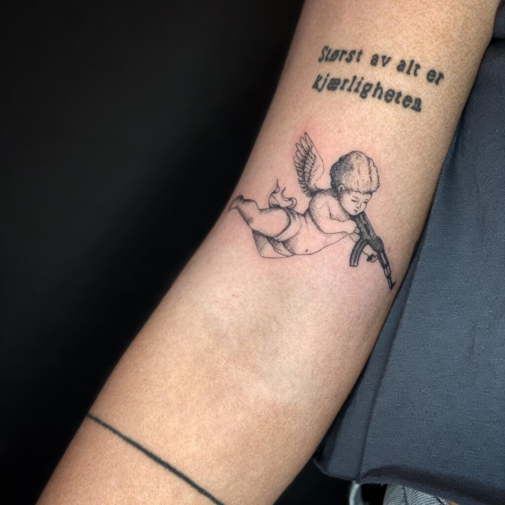 Angel with Gun Tattoo and Text Above the Elbow: Heavenly Vigilance