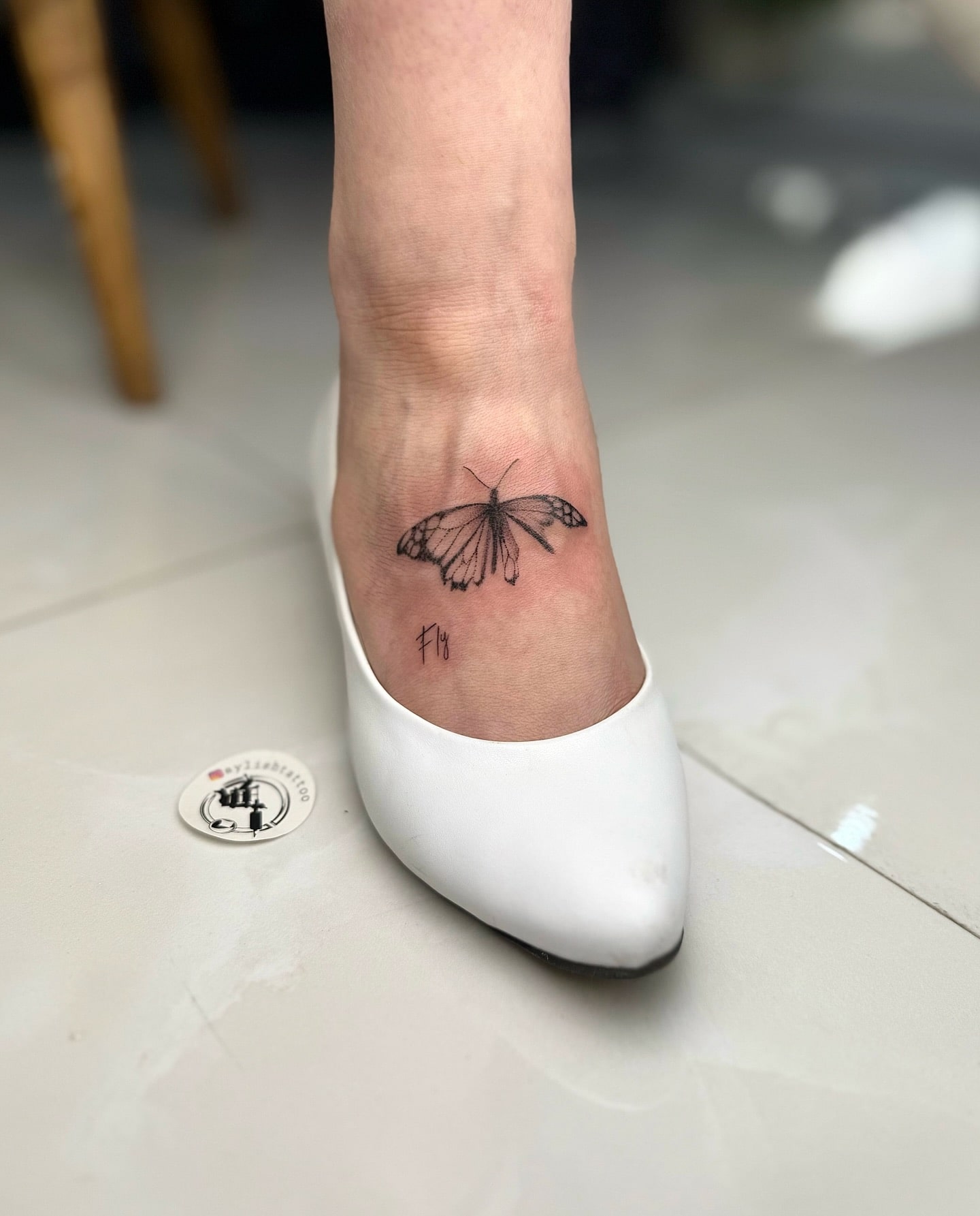 Ankle butterfly tattoo with 'Fly' text in handwritten minimalist style