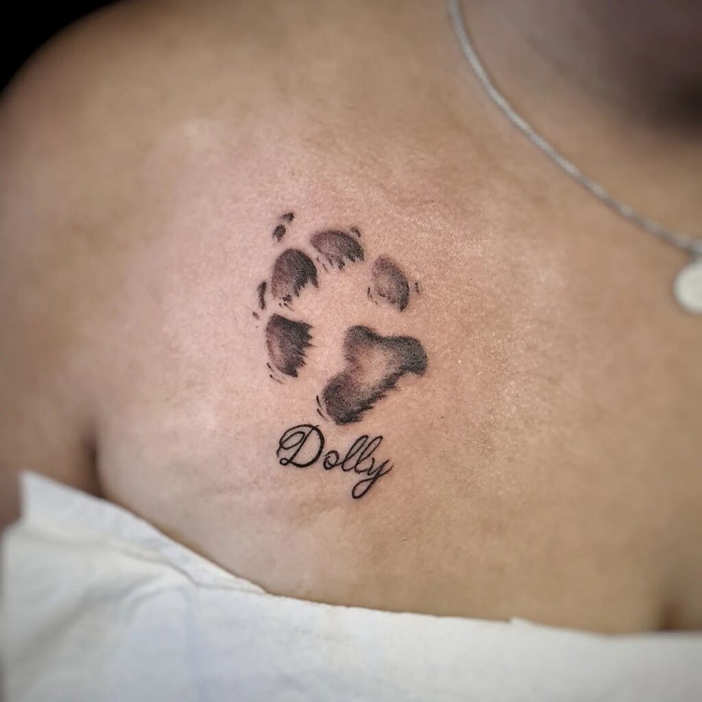 Black and Grey Dog Paw Tattoo with 'Dolly' Lettering on the Collarbone: Dolly's Tribute