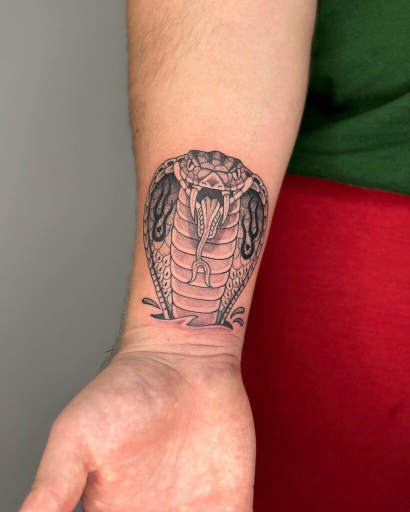 Cobra head tattoo on the inner wrist, head up and tongue out in fierce posture: angry coil