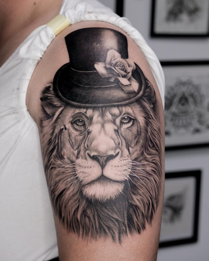 Cool lion face tattoo on the upper arm, black and grey shaded, sporting a black hat and one eyed glasses hipster king