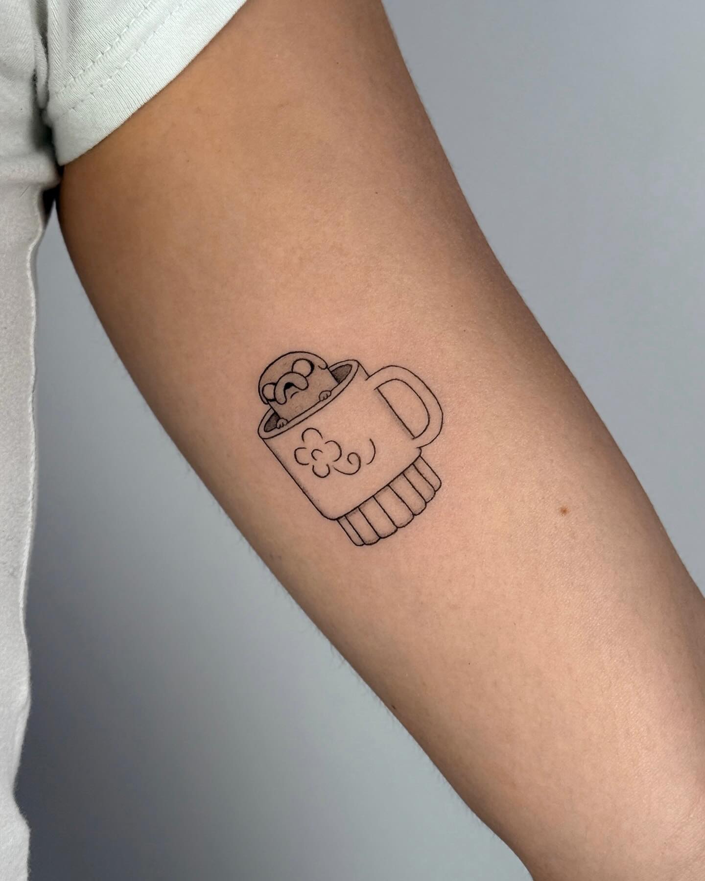 Cozy and minimalist coffee mug tattoo with a cute character peeking out, perfect for those who love comforting and whimsical tattoo designs