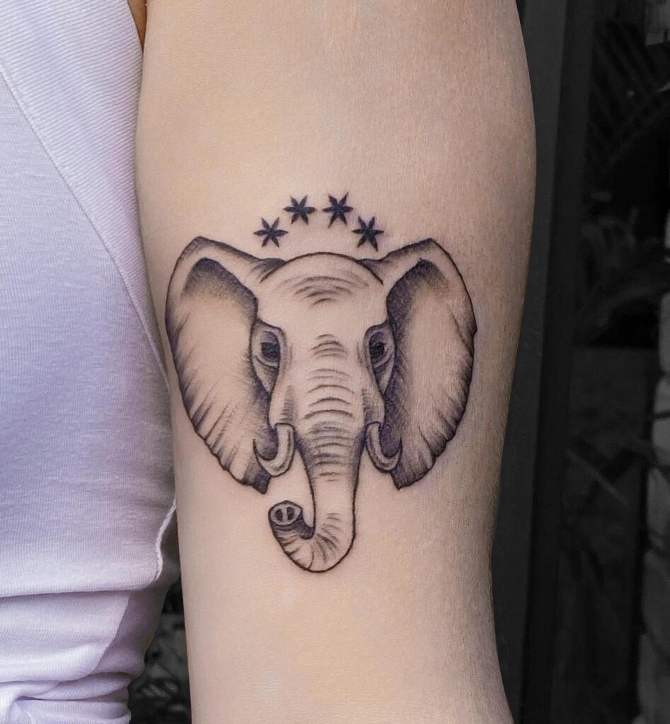 Elephant front face tattoo on forearm, trunk raised with four stars in black and grey stellar guardian