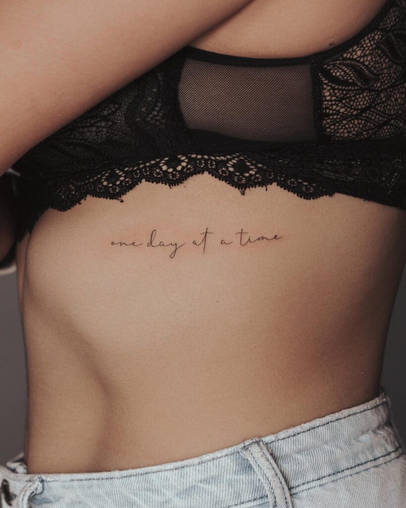 Fineline 'one day at a time' text lettering tattoo on the rib for positive encouragement daily mantra