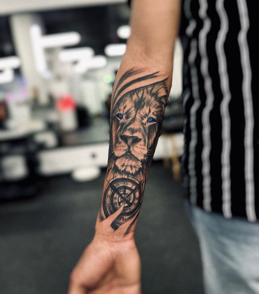 Forearm covered with lion and clock tattoo, featuring a lion with striking blue eyes above and a blackwork clock below time and roar