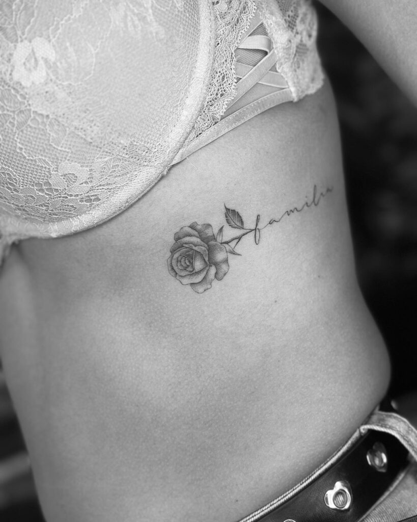 Grey shaded rose tattoo on the rib with 'feminine' script: elegant horizontal design