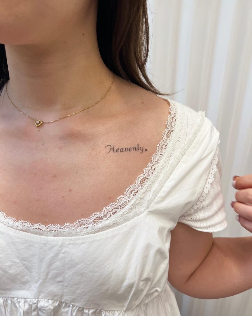 'heavenly' word lettering tattoo on collarbone with a tiny heart, subtle and sweet heavenly affirmation
