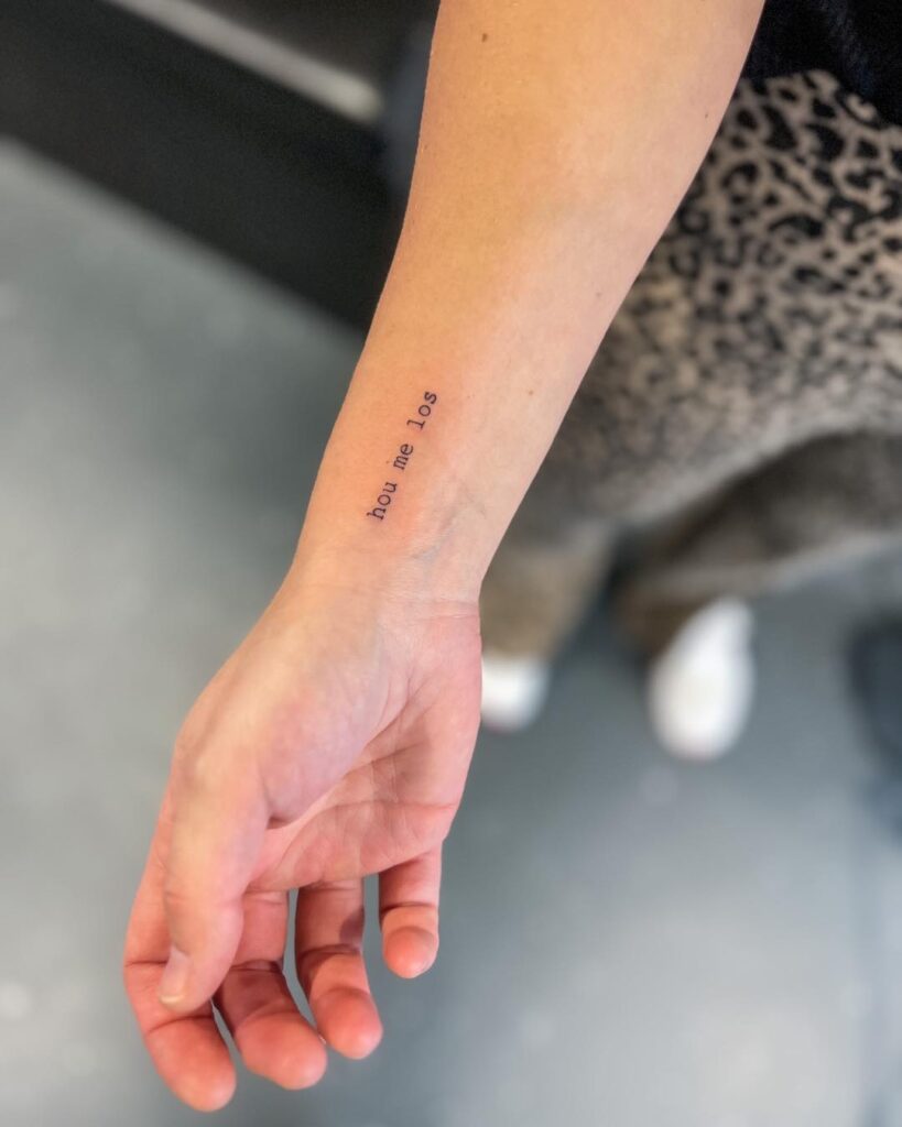 'Hou Me Los' Lettering Tattoo on the Side of the Wrist in Typewriter Font, Expressing a Sentimental Message: Hold Me Close