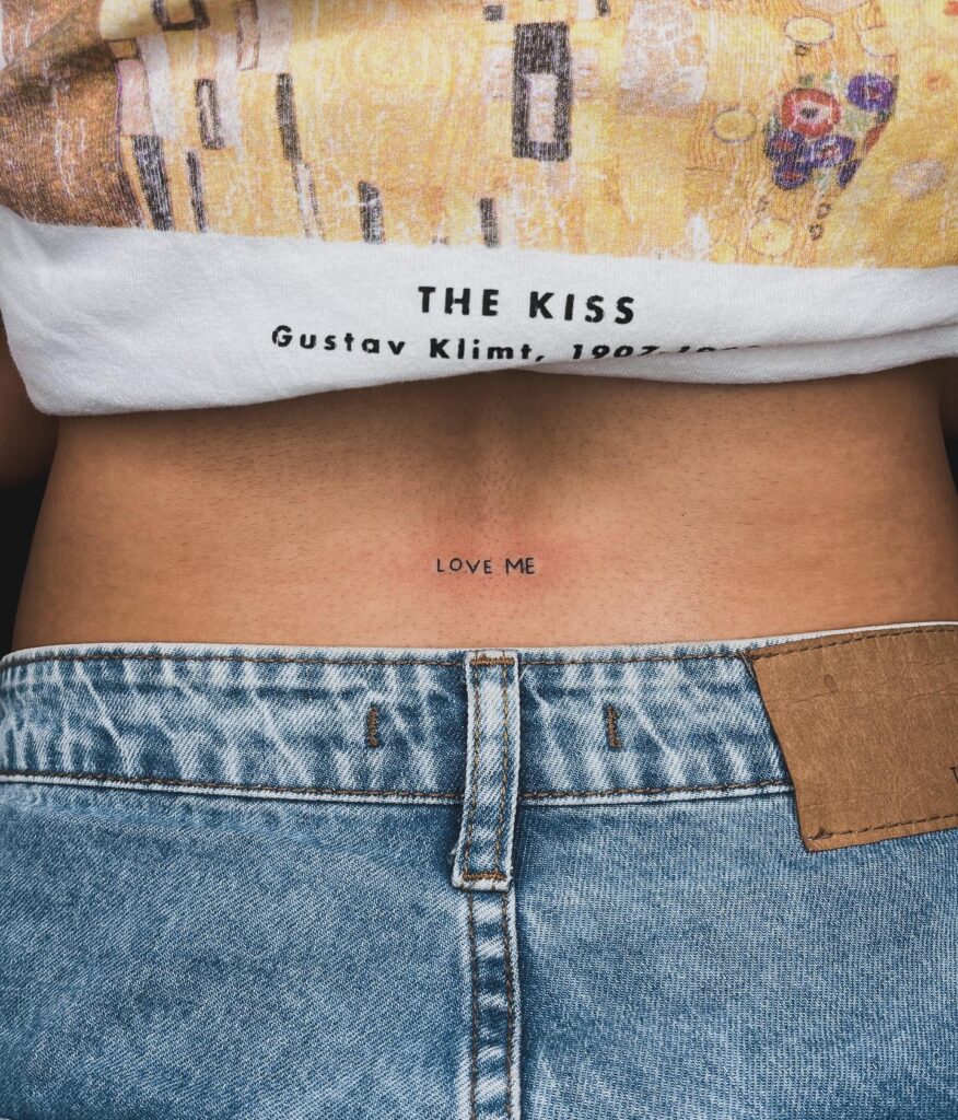 'Love me' minimal word lettering tattoo on the lower back a whisper of affection subtle plea