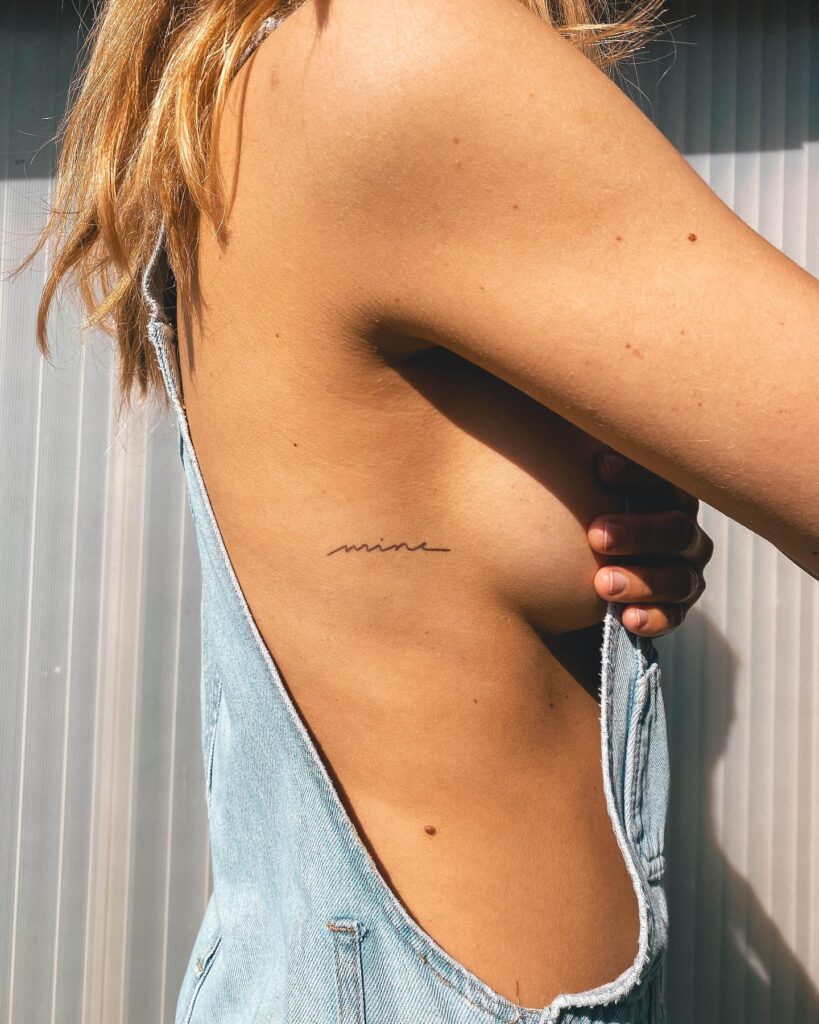 'Mine' script lettering tattoo on the side rib, a subtle and personal mark ownership ink