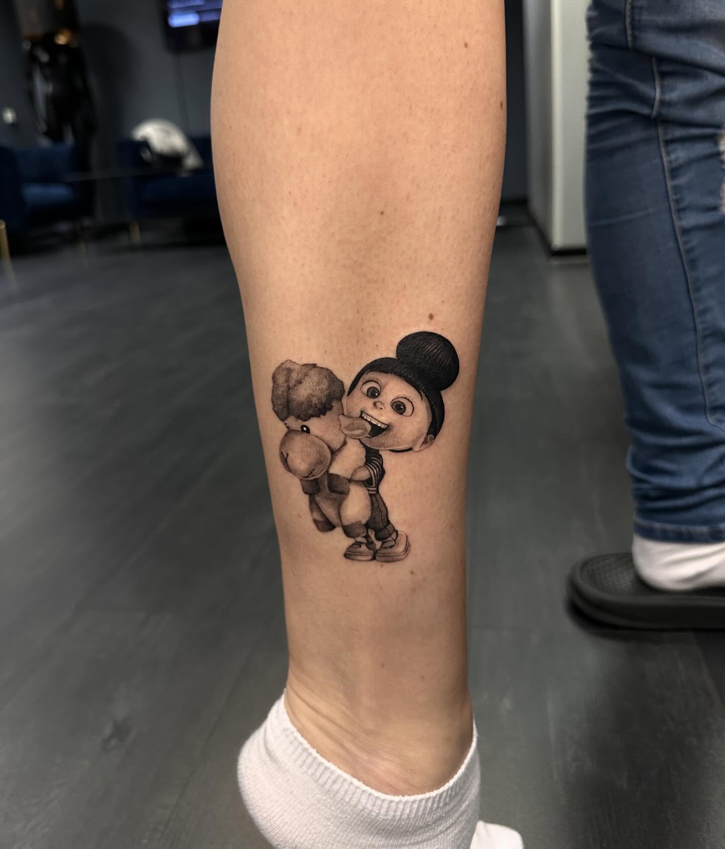 Minimalist black-and-grey Agnes tattoo hugging a fluffy stuffed animal on the lower leg, symbolizing innocence, happiness, and nostalgia