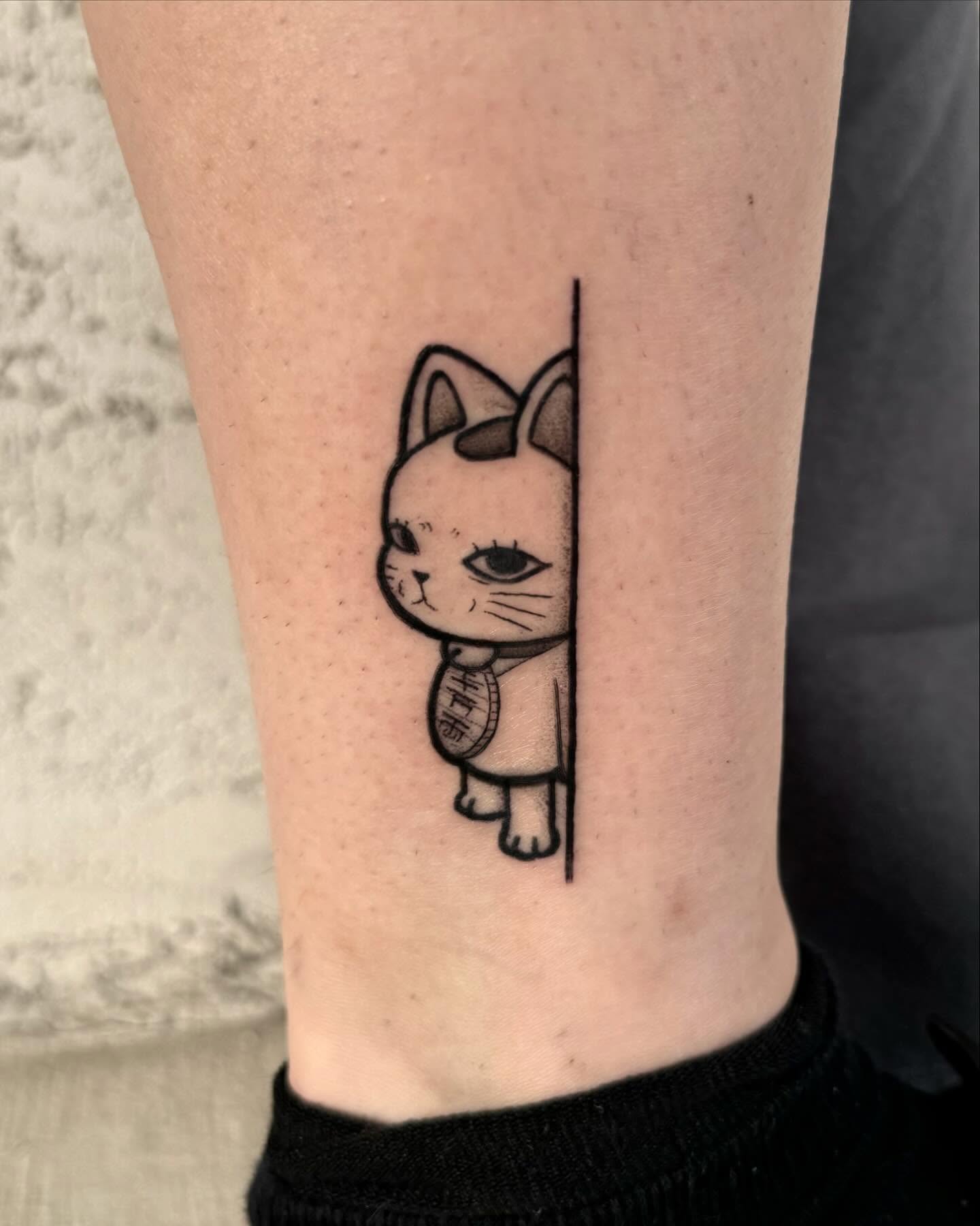 Playful fine-line cat tattoo peeking from behind a straight line, holding a lucky Maneki-Neko charm, representing curiosity, independence, and prosperity