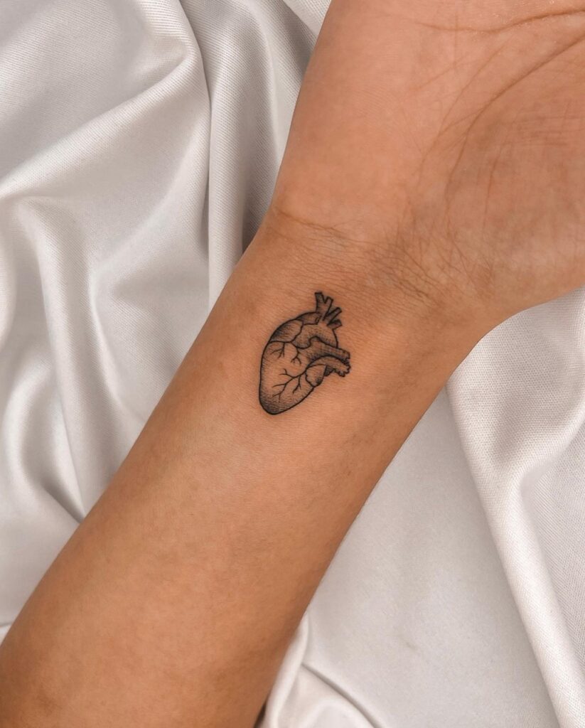 Real heart tattoo on the inner wrist, black outline with subtle grey shading heartfelt elegance