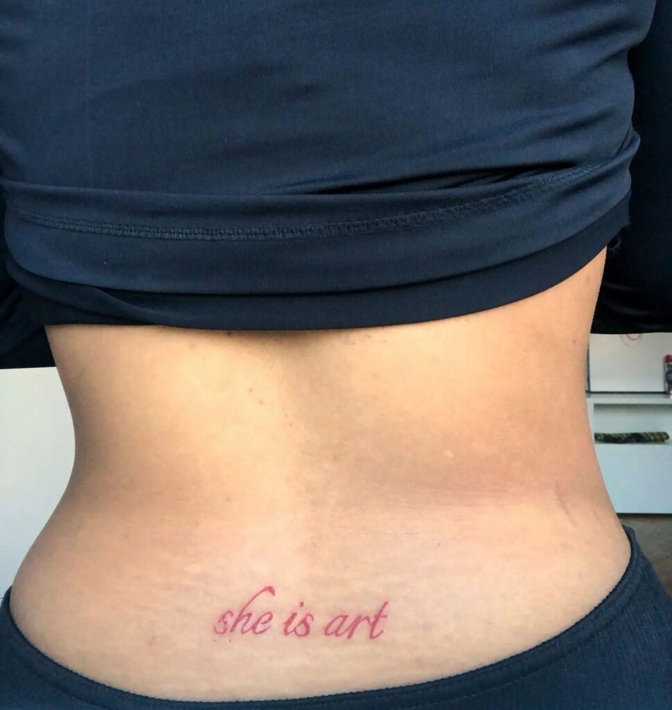 Red 'She is Art' Script Lettering Tattoo on the Lower Back: Passionate Canvas