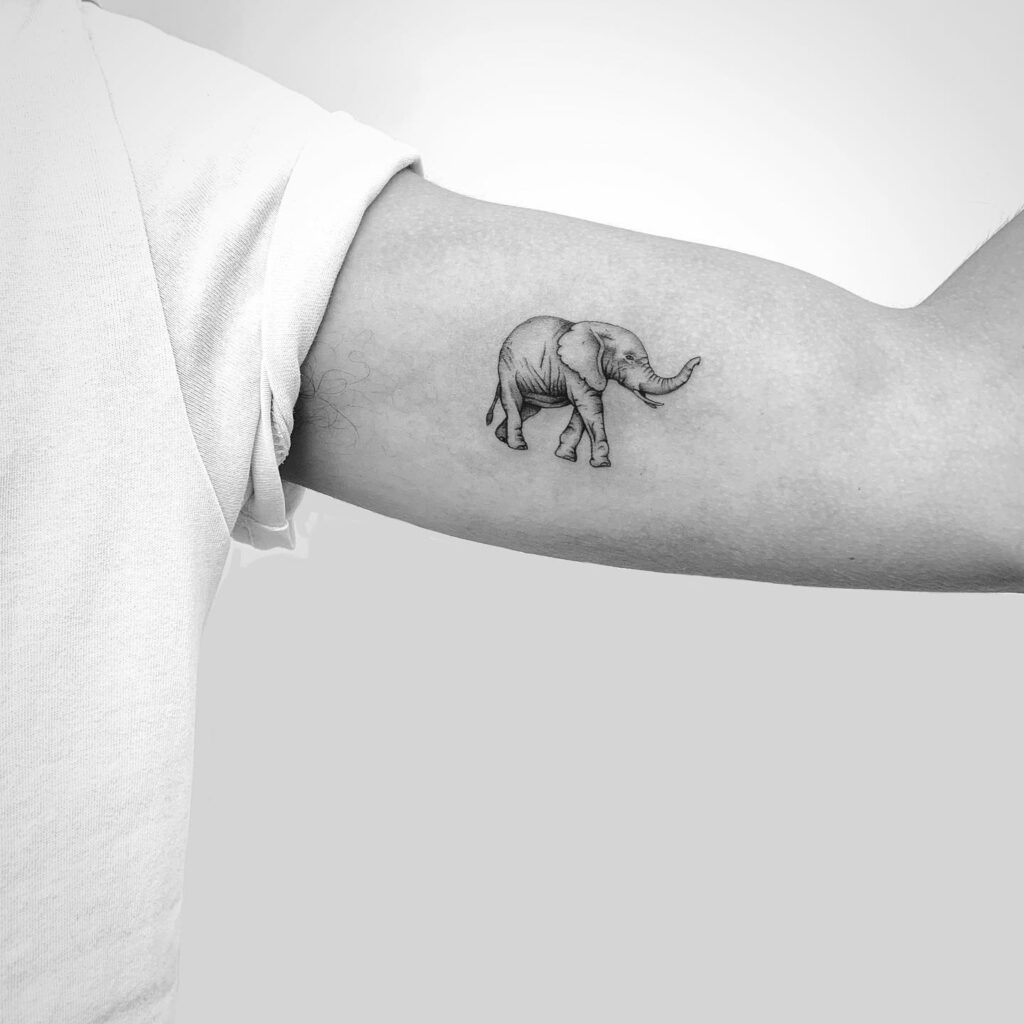 Small black and grey elephant walking with trunk lifted tattoo on bicep, grey shaded and side view unique wanderer