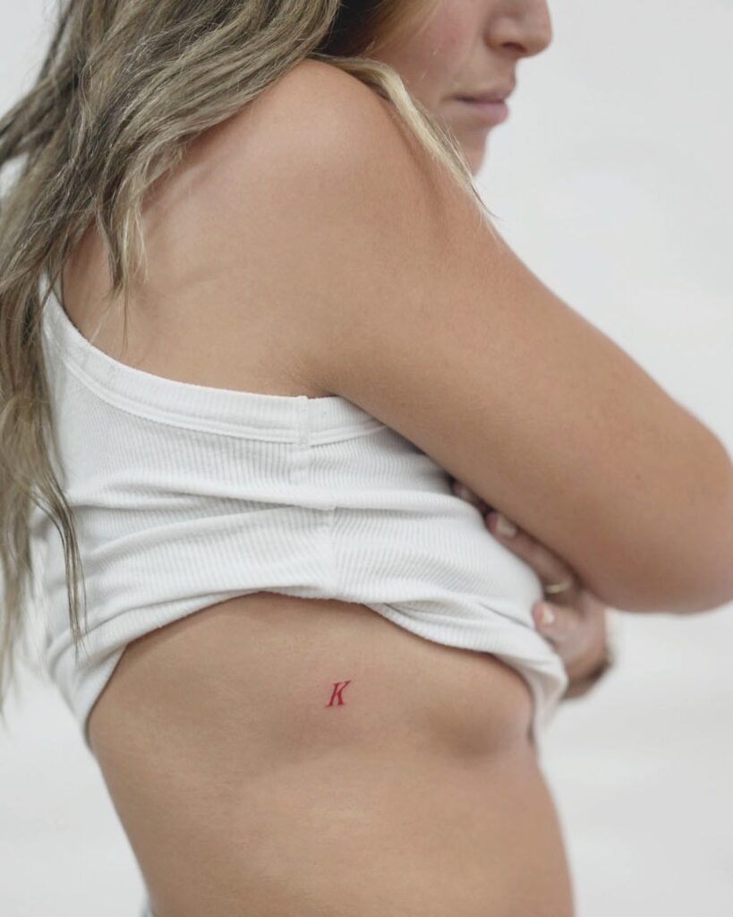 Small red 'k' letter tattoo on the side boob kissed by crimson