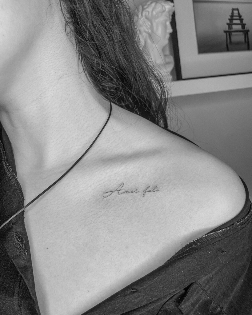 Thin-font handwriting tattoo on the collarbone: 'amor fati'