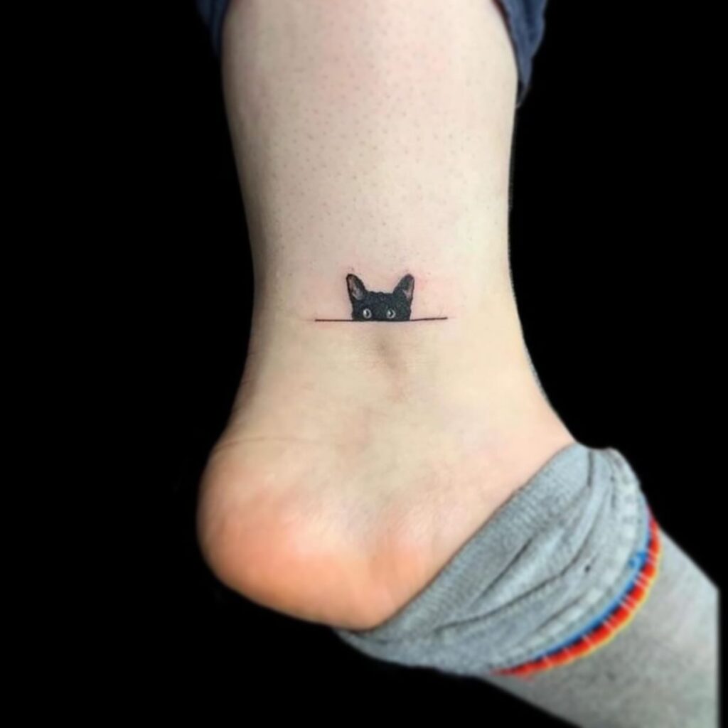 Tiny black cat ankle tattoo peeking over a line minimalist design
