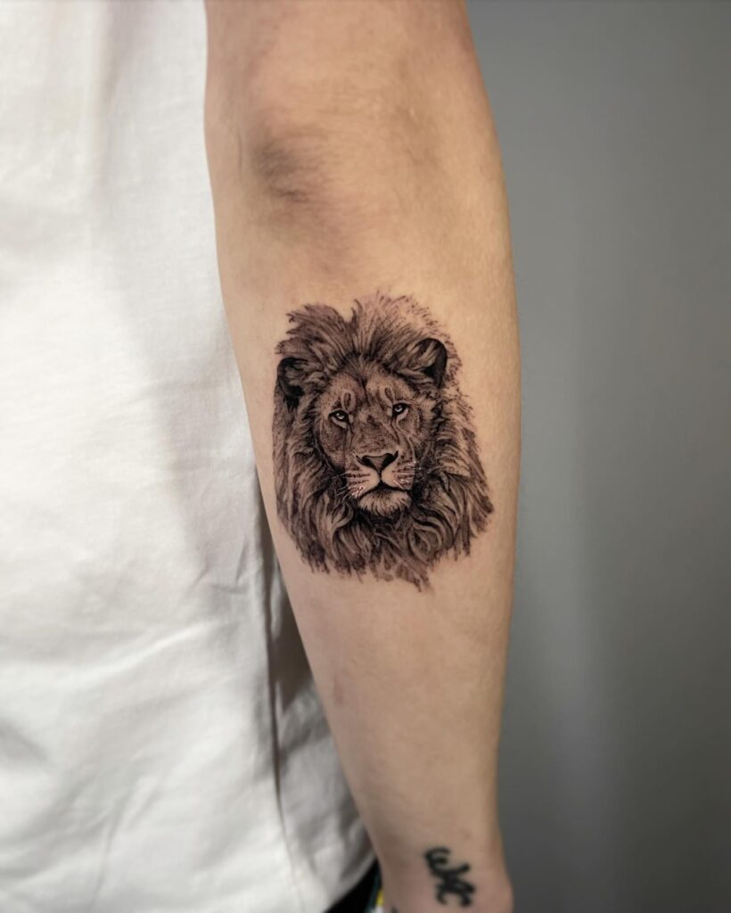 Tiny lion head tattoo on the outer forearm, black and grey shaded with exquisite detail in the mane micro majesty
