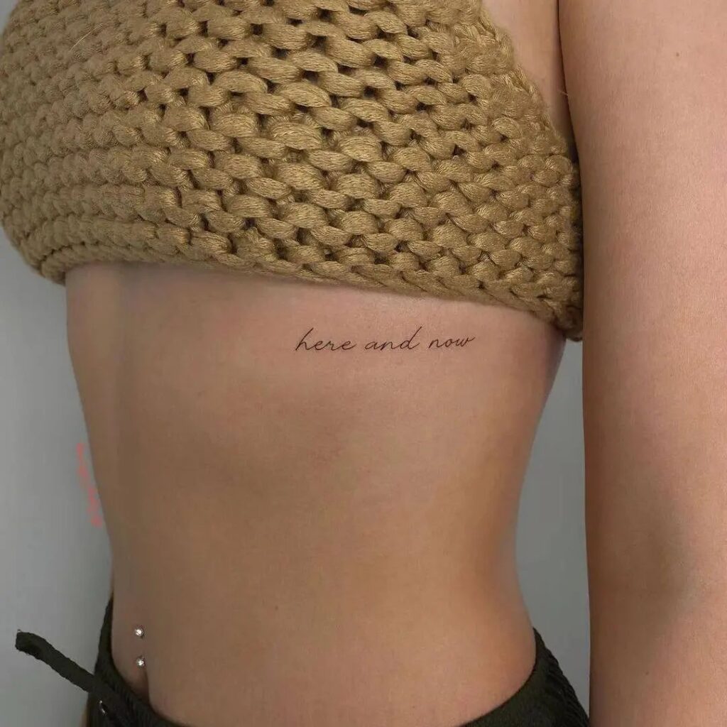 Tiny script lettering 'here and now' words rib tattoo, a subtle reminder: present moment