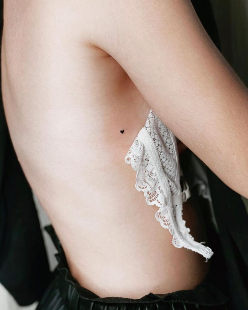 Very tiny small heart with filled black ink tattoo on the side rib for females: subtle affection