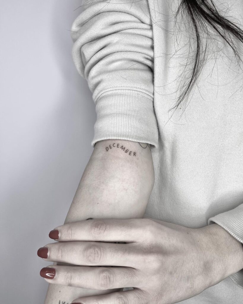 'December' Wavy Text Effect Lettering Tattoo on the Above the Elbow: Whimsical Vibes