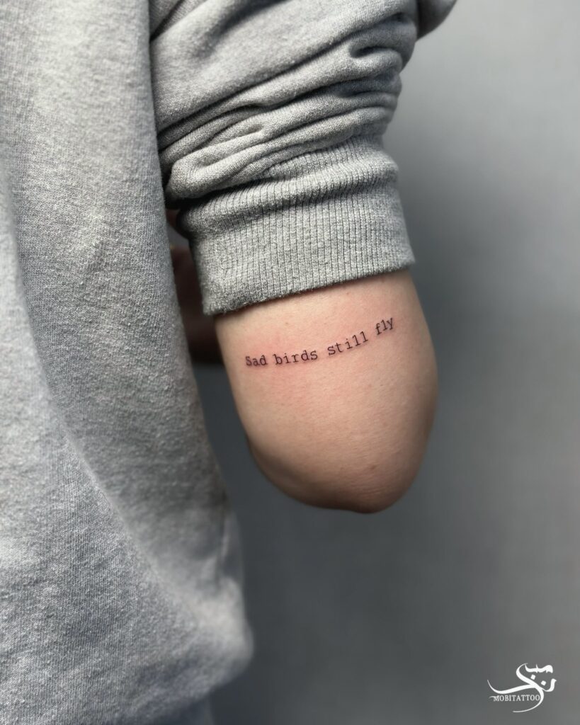 'Sad Birds Still Fly' Text Tattoo on the Back of the Above the Elbow: Melancholic Flight
