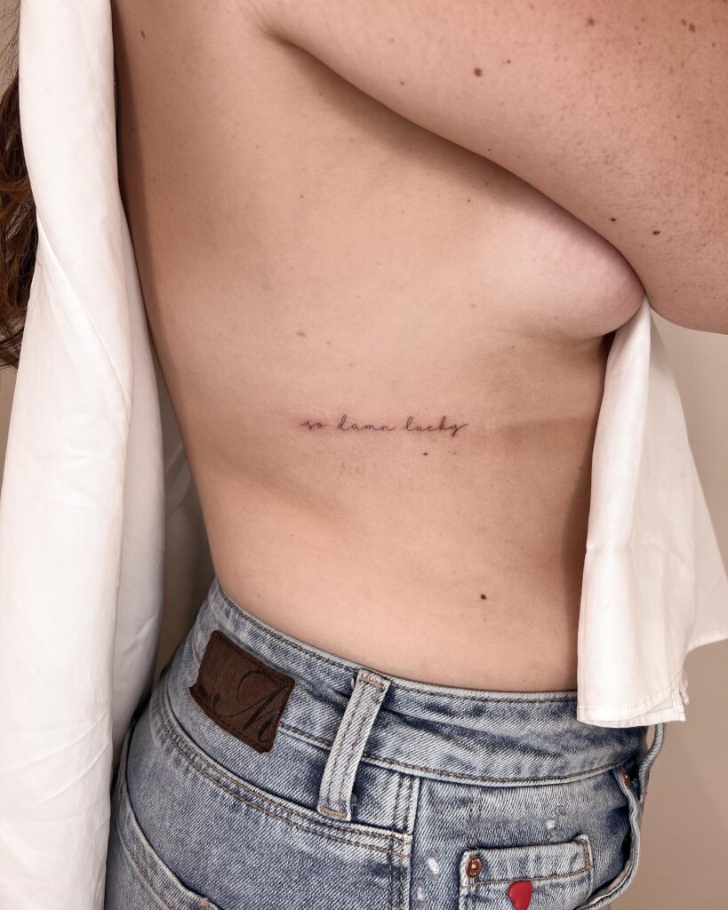 'so damn lucky' handwriting script lettering tattoo on the rib: lucky whispers